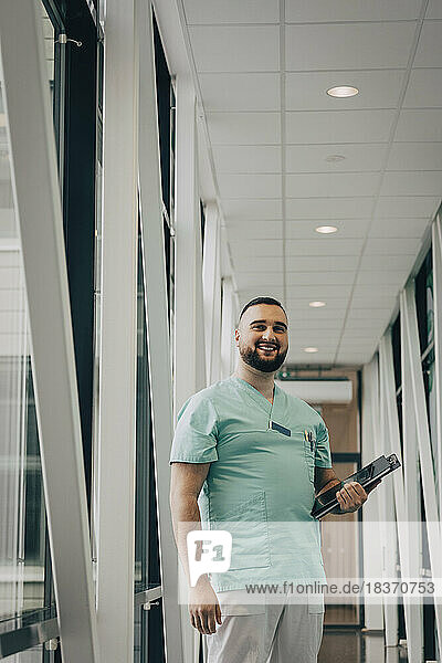 Lächelnder männlicher Mitarbeiter des Gesundheitswesens  der eine Akte hält  während er im Flur eines Krankenhauses steht