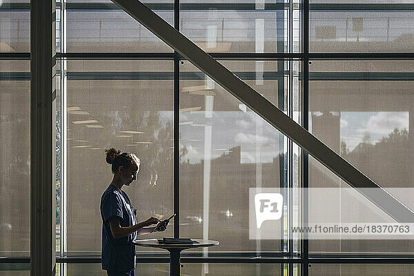 Seitenansicht einer Krankenschwester  die einen Tablet-PC benutzt  während sie am Fenster eines Krankenhauses steht