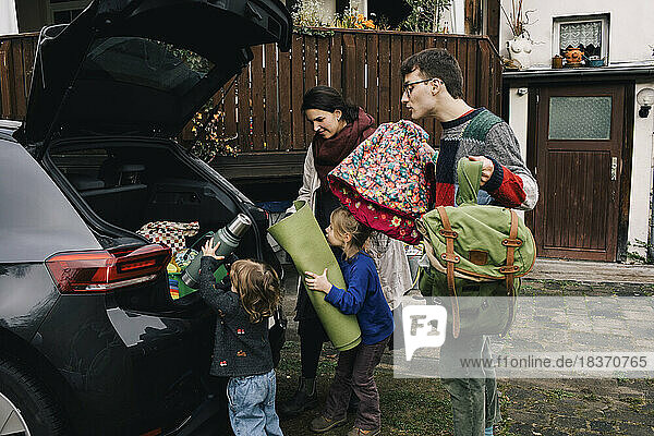 Aufgeregte Familie beim Einladen von Picknickutensilien in den Kofferraum eines Elektroautos vor dem Haus