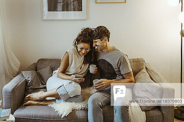 Happy couple having coffee while sharing smart phone sitting on sofa at home