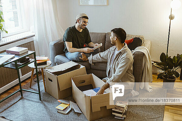 Lächelnder Mann  der auf dem Sofa sitzt und sich mit seinem Freund unterhält  während er zu Hause einen Karton auspackt