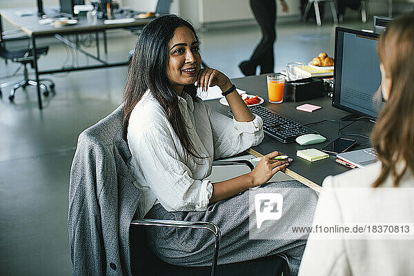 Seitenansicht einer lächelnden Geschäftsfrau im Gespräch mit einem Kollegen  während sie auf einem Stuhl im Büro sitzt