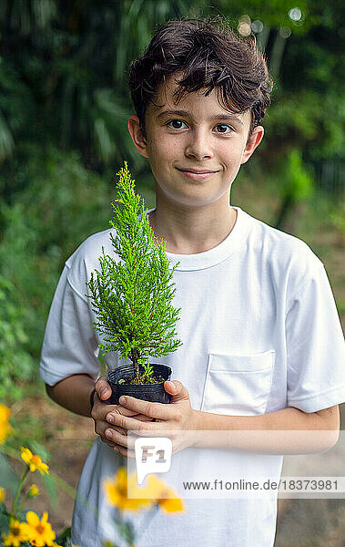 A boy holding a small tree sapling in a pot  standing in a garden.