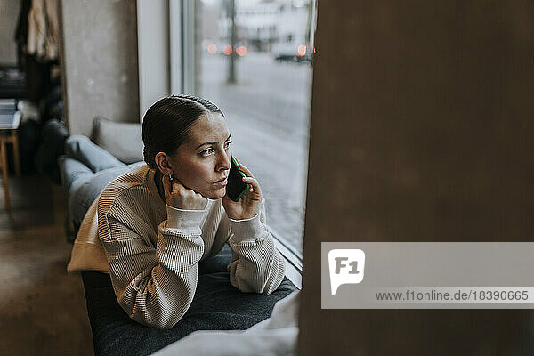 Geschäftsfrau  die über ein Mobiltelefon spricht und aus dem Fenster schaut  während sie auf dem Sitz im Büro liegt