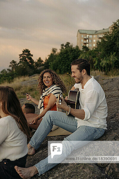 Smiling young man playing guitar while sitting with friends on rock during picnic