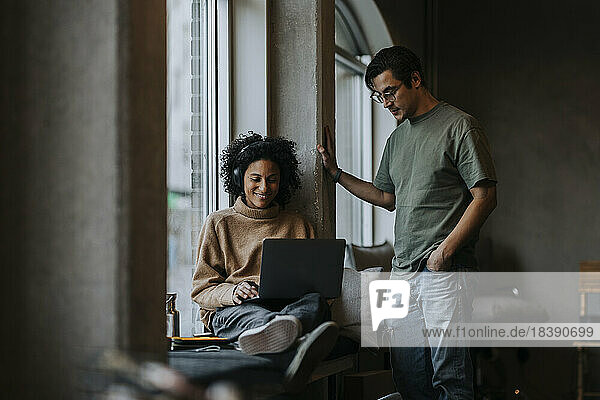 Lächelnde Geschäftsfrau  die einen Laptop benutzt  neben einem männlichen Kollegen  der in einem kreativen Büro steht