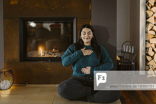 Woman with eyes closed doing breathing exercise while sitting cross-legged near fireplace at home