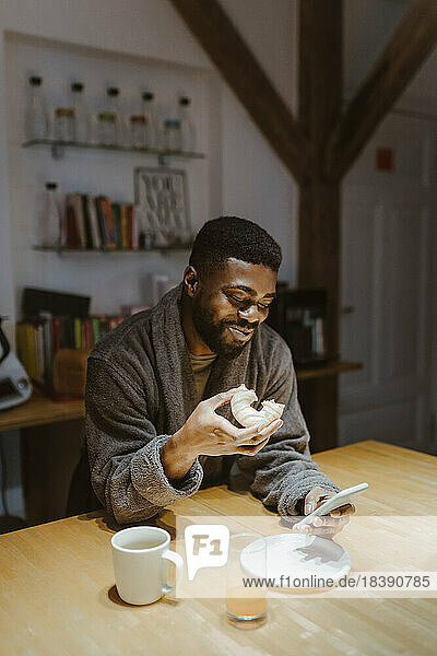 Lächelnder Mann  der sein Smartphone benutzt und zu Hause einen Doughnut isst