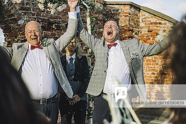 Happy gay couple holding hands while enjoying wedding ceremony by minister on sunny day