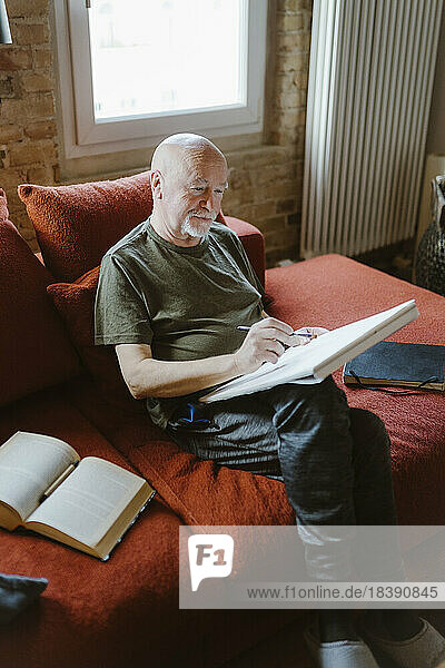 Älterer Mann beim Malen  während er zu Hause auf dem Sofa bei einem Buch sitzt
