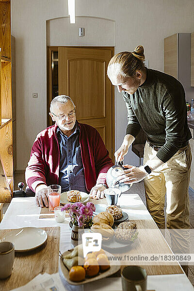 Young man pouring coffee for retired senior father with disability during breakfast at home