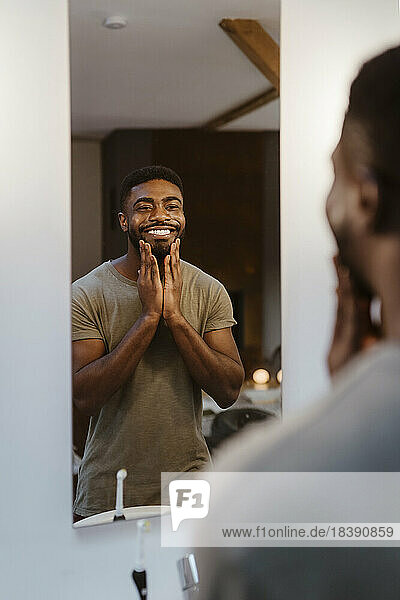 Lächelnder Mann  der Feuchtigkeitscreme aufträgt  während er sich zu Hause im Spiegel betrachtet
