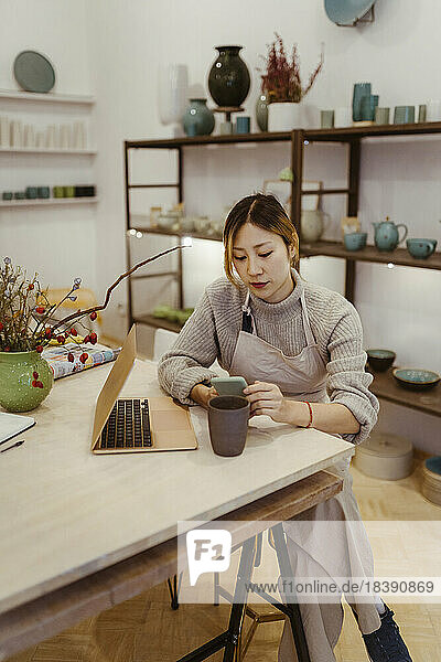 Junge Unternehmerin mit Laptop und Mobiltelefon in der Werkstatt sitzend