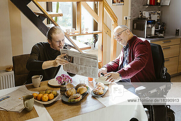 Junger Mann zeigt seinem älteren Vater mit Behinderung beim Frühstück zu Hause die Zeitung