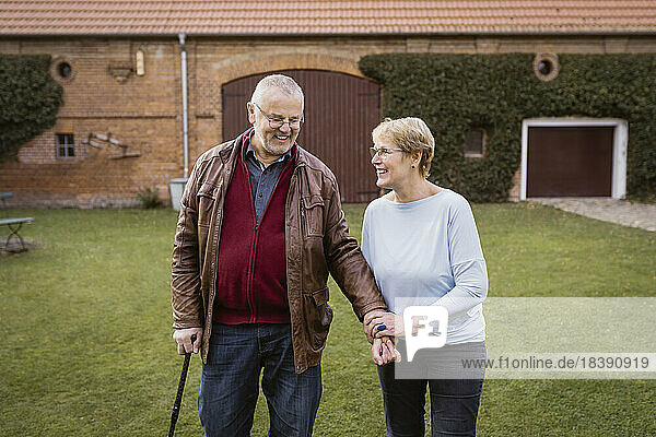 Lächelnde ältere Frau hält die Hand eines Mannes  der mit einem Stock auf einer Wiese im Hinterhof geht
