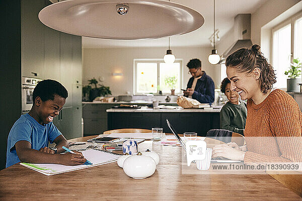 Happy mother using laptop with son coloring in book while sitting at dining table in home