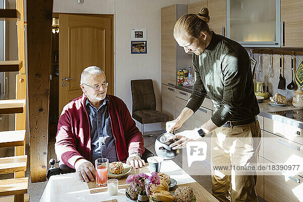 Junger männlicher Hausmeister gießt Kaffee für einen älteren Mann im Ruhestand mit Behinderung beim Frühstück zu Hause ein