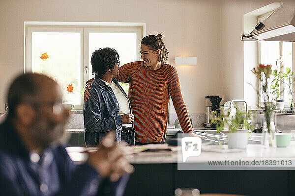 Happy daughter-in-law with arm around senior woman standing in kitchen at home