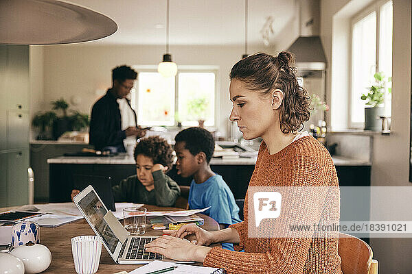 Seitenansicht einer Frau  die einen Laptop benutzt  während die Familie im Hintergrund zu Hause ist