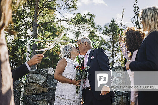 Senior bride with bouquet kissing groom on mouth amidst guest during wedding celebration