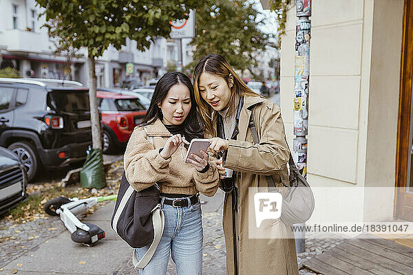 Young female friends sharing smart phone while standing on sidewalk