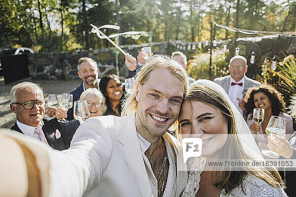 Porträt eines glücklichen frisch vermählten Paares  das am Hochzeitstag ein Selfie mit Gästen im Hintergrund macht