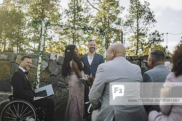 Glückliches frisch vermähltes Paar  das einen Blumenstrauß hält  umgeben von Gästen und einem behinderten Pfarrer bei der Hochzeitszeremonie
