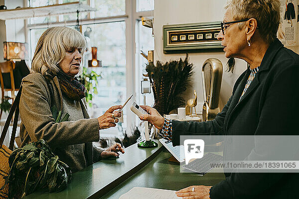 Ältere Kundin im Gespräch mit dem Besitzer  während sie in einem Geschäft online bezahlt