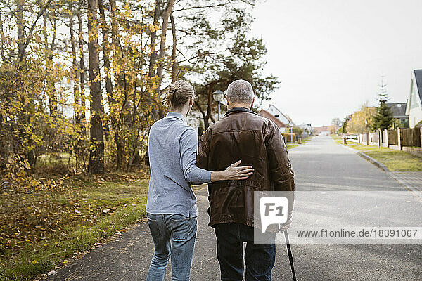 Rückansicht eines Hausmeisters  der einen älteren Mann im Ruhestand unterstützt  der mit einem Stock auf der Straße geht
