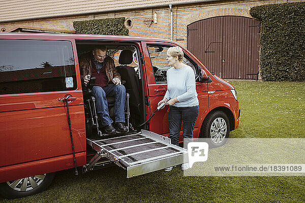 Ältere Frau hält die Fernbedienung in der Hand  während sie einem Mann mit Behinderung im motorisierten Rollstuhl beim Aussteigen aus einem Lieferwagen hilft