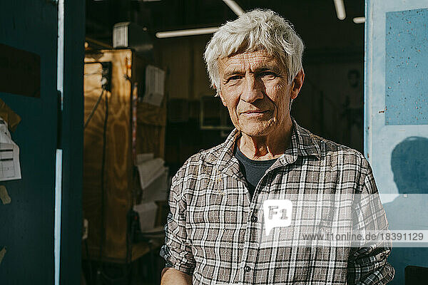 Porträt eines älteren männlichen Schreiners in der Nähe der Werkstatttür an einem sonnigen Tag