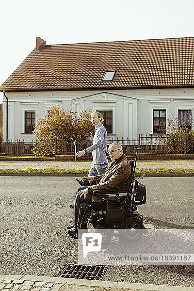 Seitenansicht eines älteren Mannes mit Behinderung in einem motorisierten Rollstuhl  der von einer jungen Betreuerin auf einer Straße an einem sonnigen Tag begleitet wird
