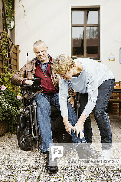Ältere Frau in voller Länge  die einem Mann mit Behinderung hilft  der in einem motorisierten Rollstuhl im Hinterhof sitzt