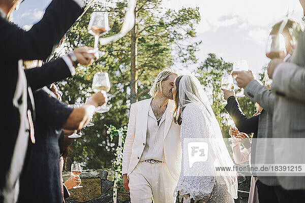 Zärtliche Braut und Bräutigam küssen sich auf den Mund inmitten von Gästen  die bei der Hochzeitszeremonie anstoßen