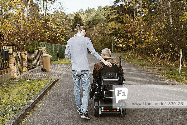 Junge männliche Pflegekraft in voller Länge  die an einem älteren Mann mit Behinderung im motorisierten Rollstuhl auf der Straße vorbeigeht
