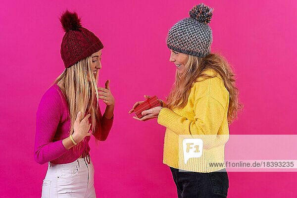 Zwei junge blonde kaukasische Frauen  die sich gegenseitig ein Geschenk überreichen  vor einem rosa Hintergrund