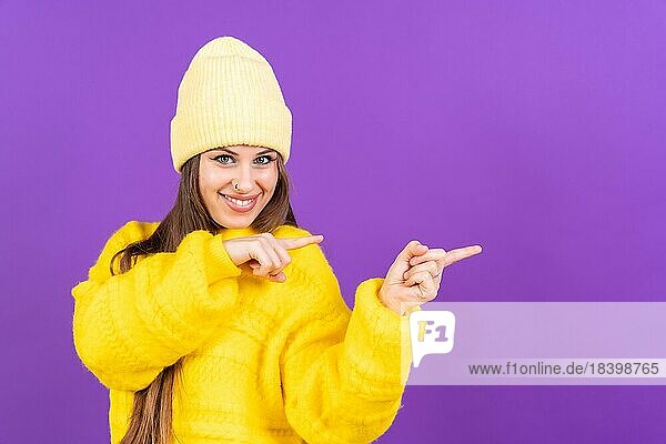 Attraktive Frau  die lächelnd mit den Fingern auf den Kopierraum auf lila Hintergrund zeigt  gelber Wollpullover