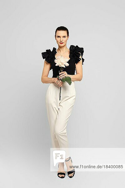 Schöne erwachsene Frau in schwarzer Rüschenbluse und weißer Hose  die elegant mit einer Blume in der Hand posiert und direkt in die Kamera schaut  in einer Innenaufnahme im Studio