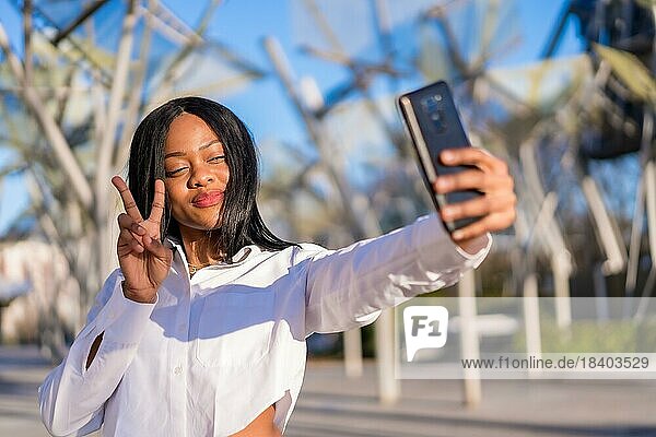 Young african woman in white clothes in the city at sunset  video call  making a victory gesture