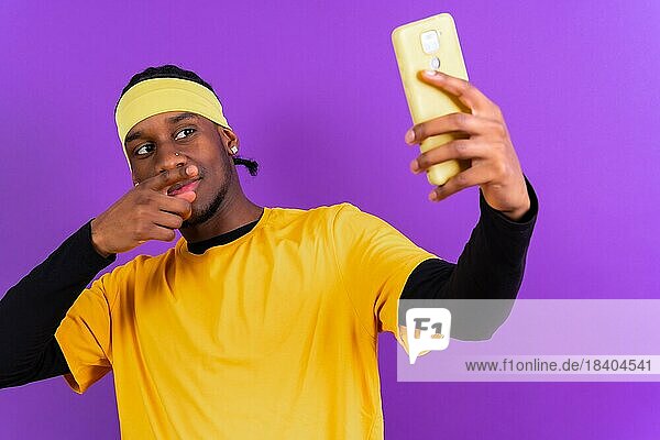 Schwarzer ethnischer Mann mit einem Telefon in gelber Kleidung auf einem lila Hintergrund  der ein Selfie macht