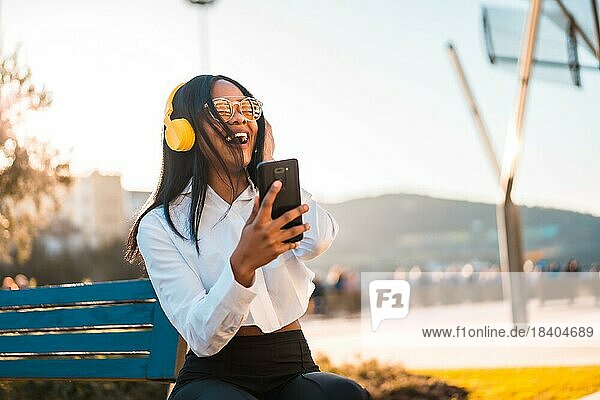 Afroamerikanische Frau hört Musik und lächelt mit Kopfhörern im Sommer bei Sonnenuntergang in der Stadt