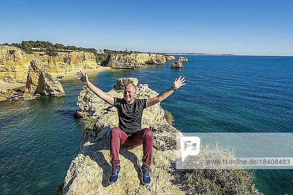 Happy man sitting with hands up on the top of cliffs at Marinha beach  Algarve  Portugal  Europe