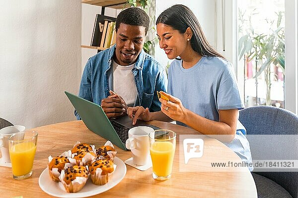 Multi-ethnic couple making purchase online with the computer while having breakfast  next to the window