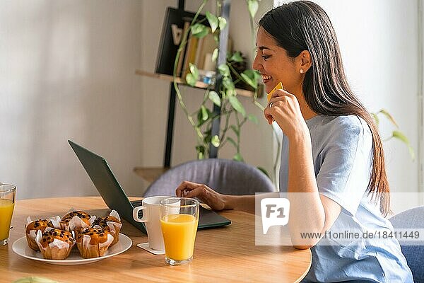 Frau  die am Computer online einkauft  während sie am Morgen am Fenster frühstückt