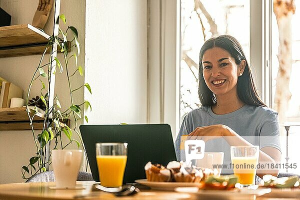 Frau beim Onlineshopping mit Computer  während sie morgens am Fenster mit Orangensaft frühstückt