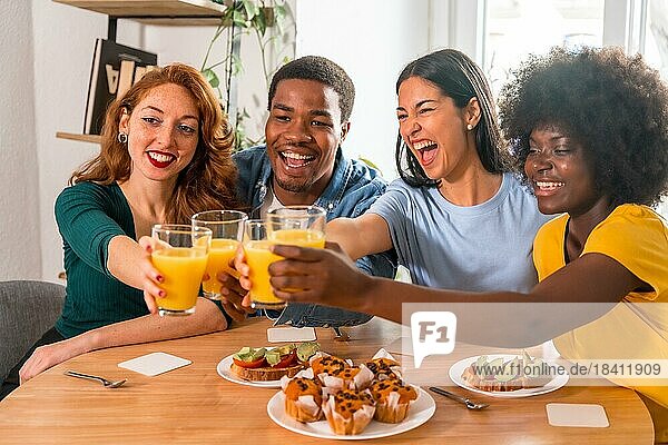 Multiethnische Freunde stoßen bei einem Frühstück mit Orangensaft und Muffins zu Hause an