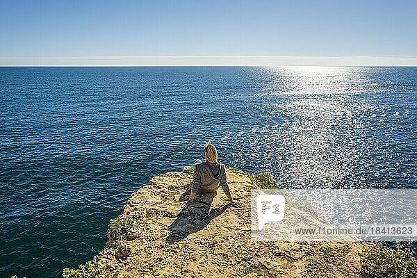 Rückenansicht einer jungen Frau  die auf den Klippen sitzt und die Schönheit des Atlantischen Ozeans am Strand von Marinha in der Algarve  Portugal  genießt  Europa