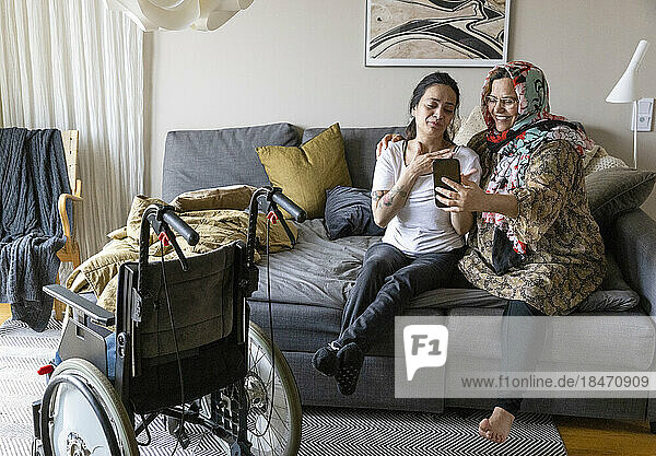 Frau mit Querschnittslähmung führt Videogespräch über ihr Smartphone  während sie mit ihrer Mutter auf dem Sofa zu Hause sitzt