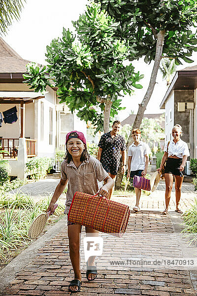 Glückliches Mädchen  das eine Matte hält  während es mit seiner Familie auf dem Fußweg in einem Touristenort spazieren geht