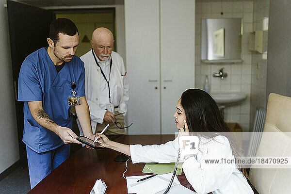 Weiblicher Arzt  der über das Telefon spricht  während er auf einem digitalen Tablet unterschreibt  das von einer Krankenschwester im Büro eines Krankenhauses gehalten wird